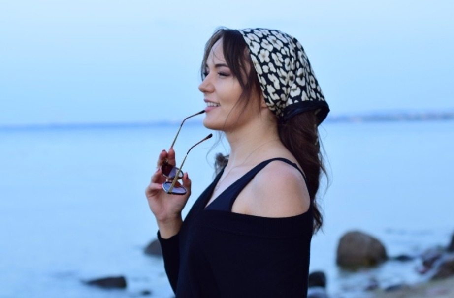 Uśmiechnięta brunetka w chustce w panterkę, trzymająca okulary przeciwsłoneczne na tle błękitnego morza.