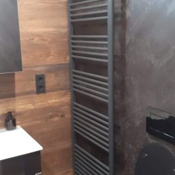 BUD-MILK MARCIN MILKE - Szary grzejnik drabinkowy łazienkowy na tle ściany z efektem betonu i płytek imitujących drewno, obok umywalka z szafką w ciemnym kolorze.