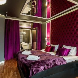 BUD-MILK MARCIN MILKE - Luksusowa sypialnia z bordowym pikowanym zagłówkiem, fioletowymi zasłonami i widokiem na łazienkę z wolnostojącą wanną, odbicie łazienki w lustrzanym suficie.