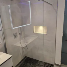 Nowoczesna kabina prysznicowa walk-in z szarymi płytkami imitującymi beton, podświetleniem LED w suficie na fioletowo oraz odpływem liniowym wykończonym czarnymi kamieniami.