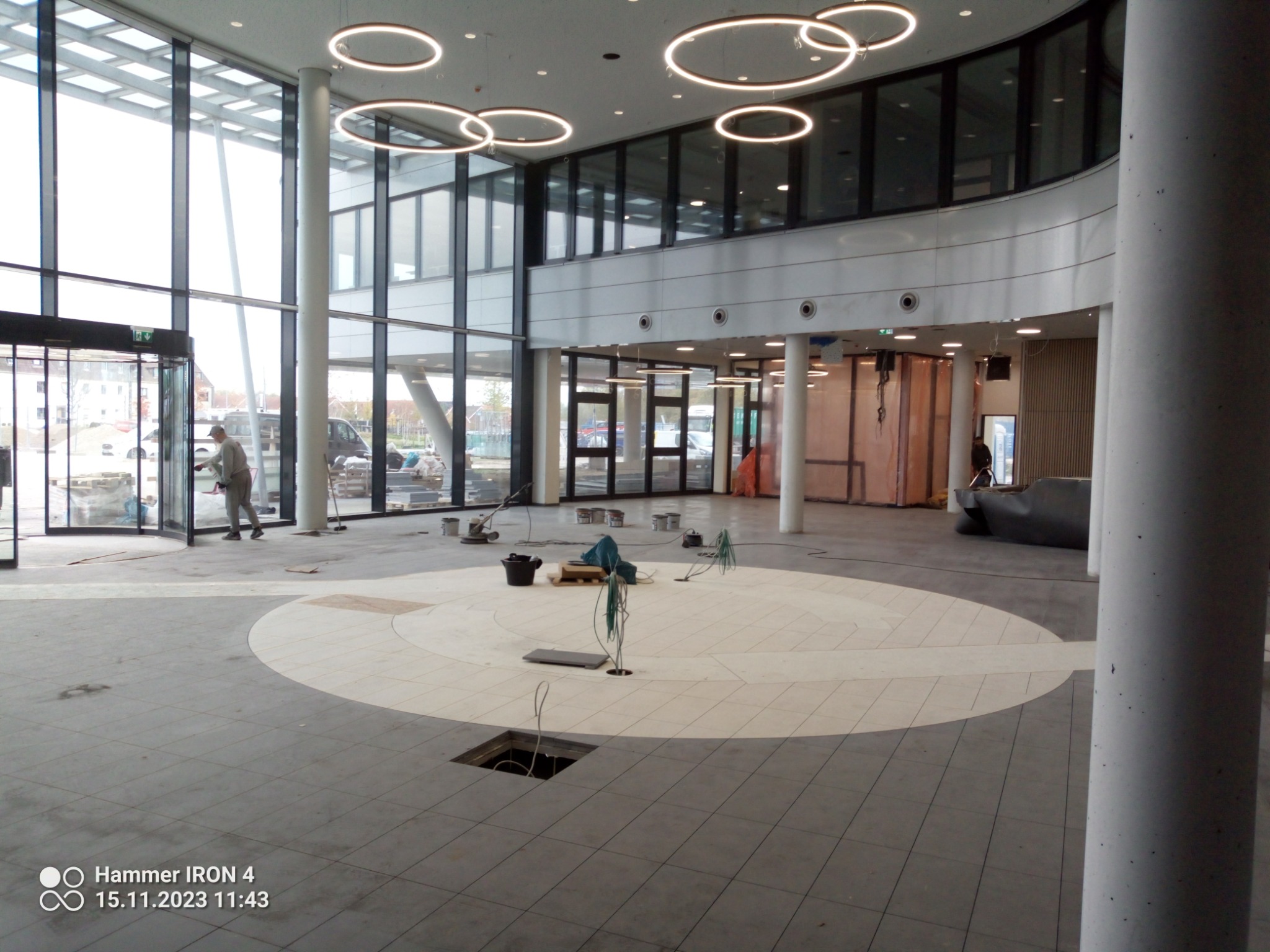 Układanie jasnych płytek w okrągły wzór na podłodze w dużym, jasnym holu, widoczne narzędzia i pracownik przy wejściu.