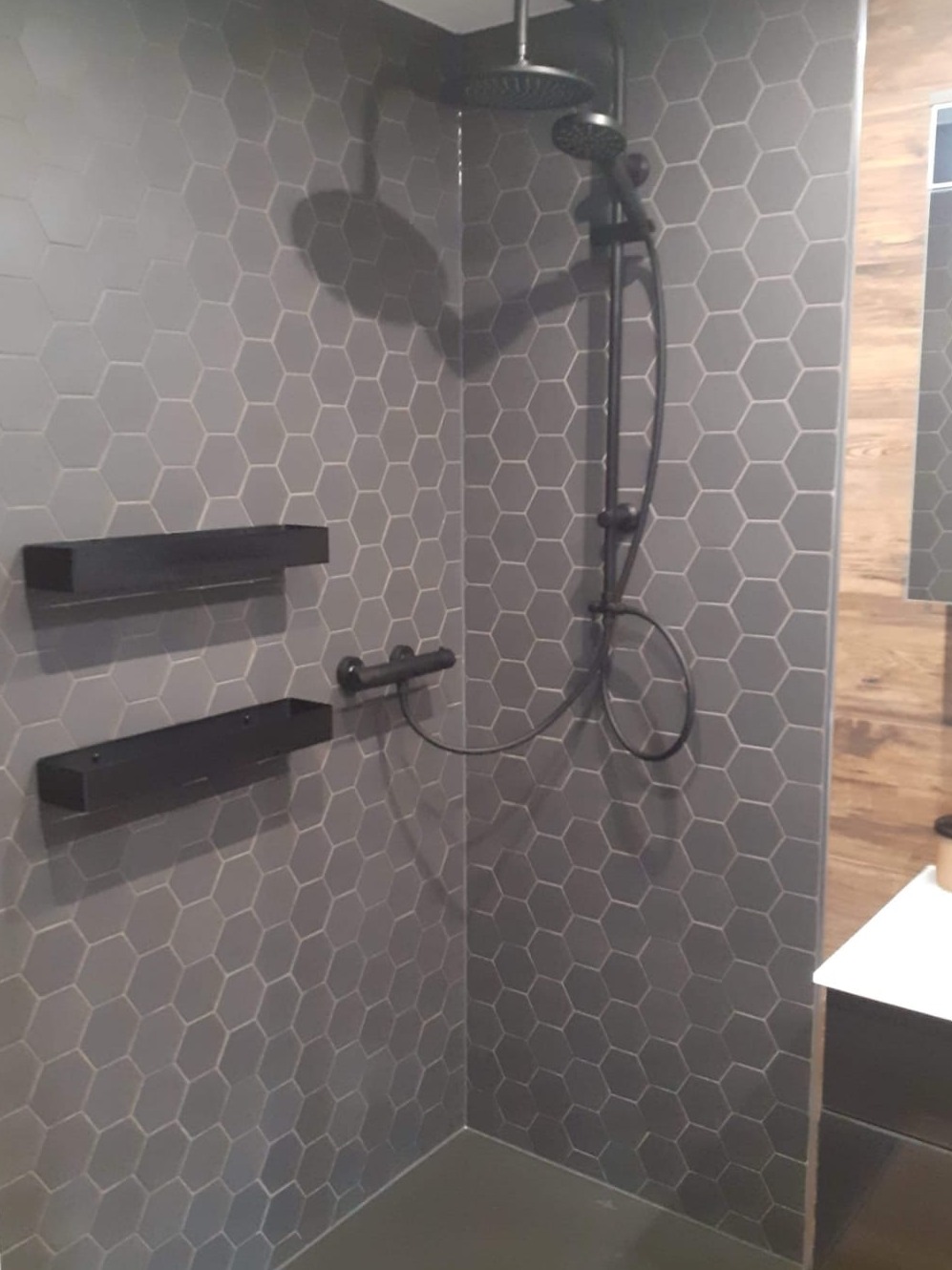 Szara kabina prysznicowa z heksagonalnymi płytkami, czarną armaturą i półkami, wykończona drewnopodobnymi panelami.