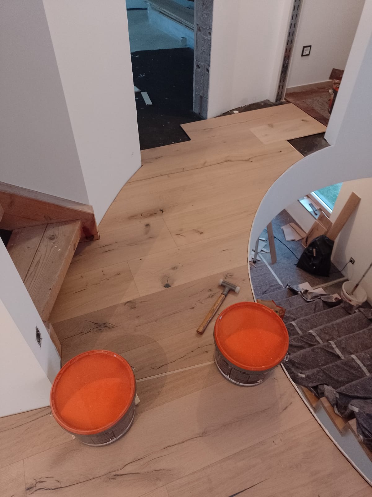 Układanie jasnych paneli podłogowych w zakrzywionym korytarzu, widoczne dwa pomarańczowe wiadra i młotek, częściowo wykończone schody drewniane i przygotowane podłoże w sąsiednim pomieszczeniu.