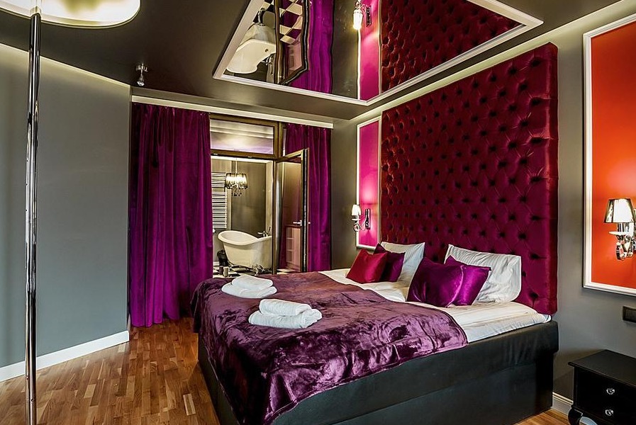 Luksusowa sypialnia z bordowym pikowanym zagłówkiem, fioletowymi zasłonami i widokiem na łazienkę z wolnostojącą wanną, odbicie łazienki w lustrzanym suficie.