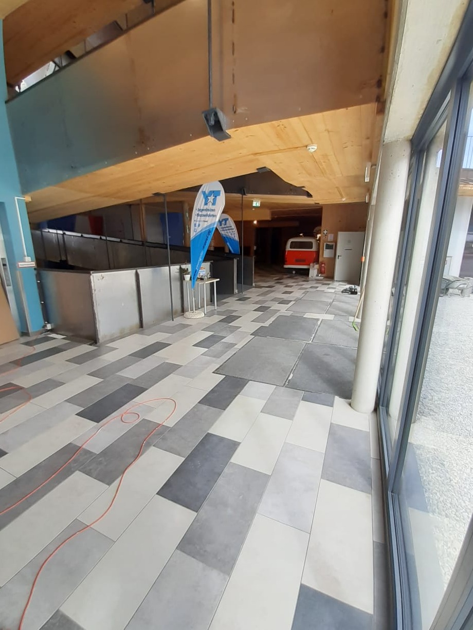 Wnętrze budynku z ułożoną podłogą z płytek w odcieniach szarości, widoczne dwa flagi reklamowe i czerwony bus w głębi korytarza.