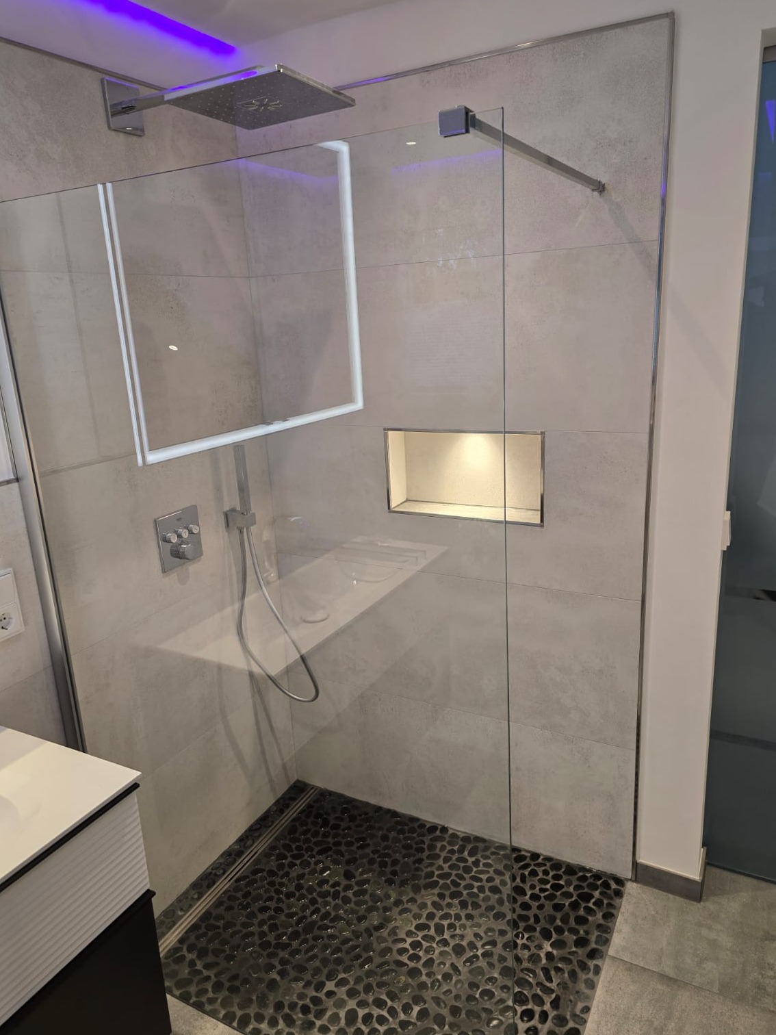 Nowoczesna kabina prysznicowa walk-in z szarymi płytkami imitującymi beton, podświetleniem LED w suficie na fioletowo oraz odpływem liniowym wykończonym czarnymi kamieniami.