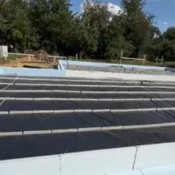 Fundamenty domu w trakcie izolacji: białe płyty styropianowe i czarna folia izolacyjna na tle zieleni i błękitnego nieba.