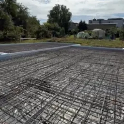 Zbrojenie płyty fundamentowej na folii izolacyjnej, widok z góry, w tle drzewa i zabudowania. Przygotowanie do wylewki betonowej, słoneczny dzień.