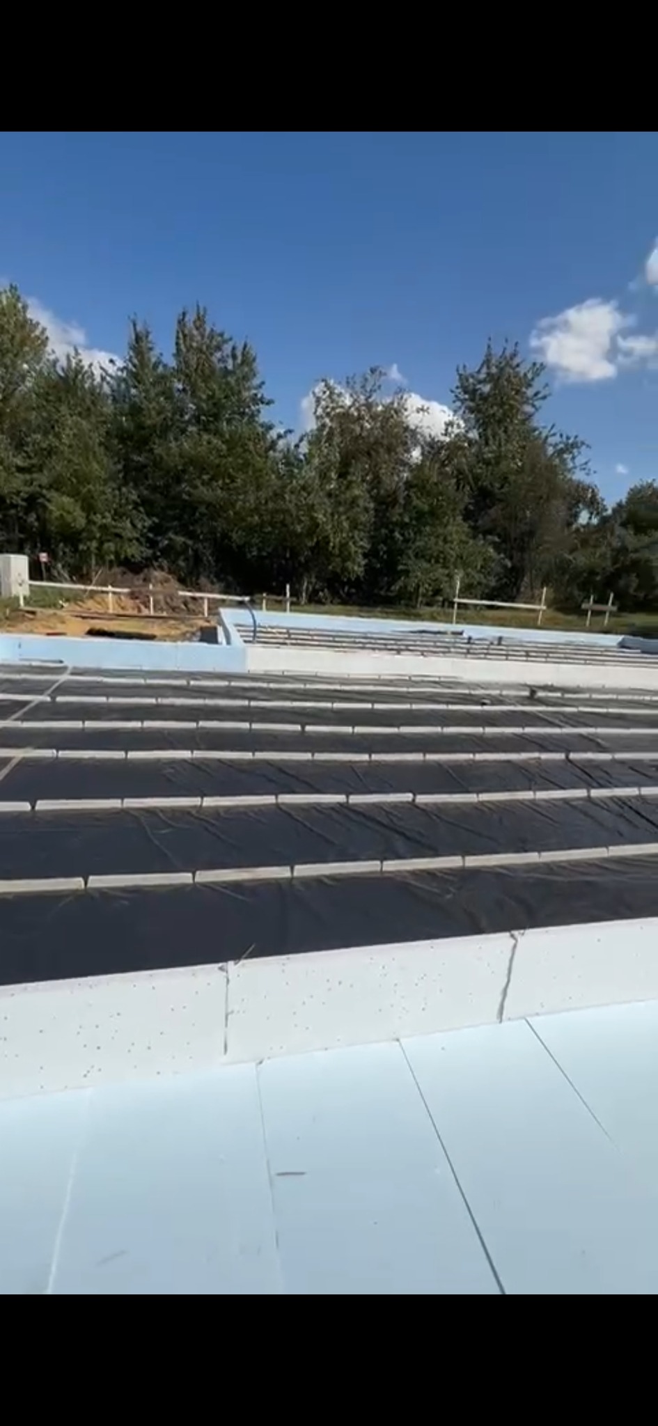 Fundamenty domu w trakcie izolacji: białe płyty styropianowe i czarna folia izolacyjna na tle zieleni i błękitnego nieba.