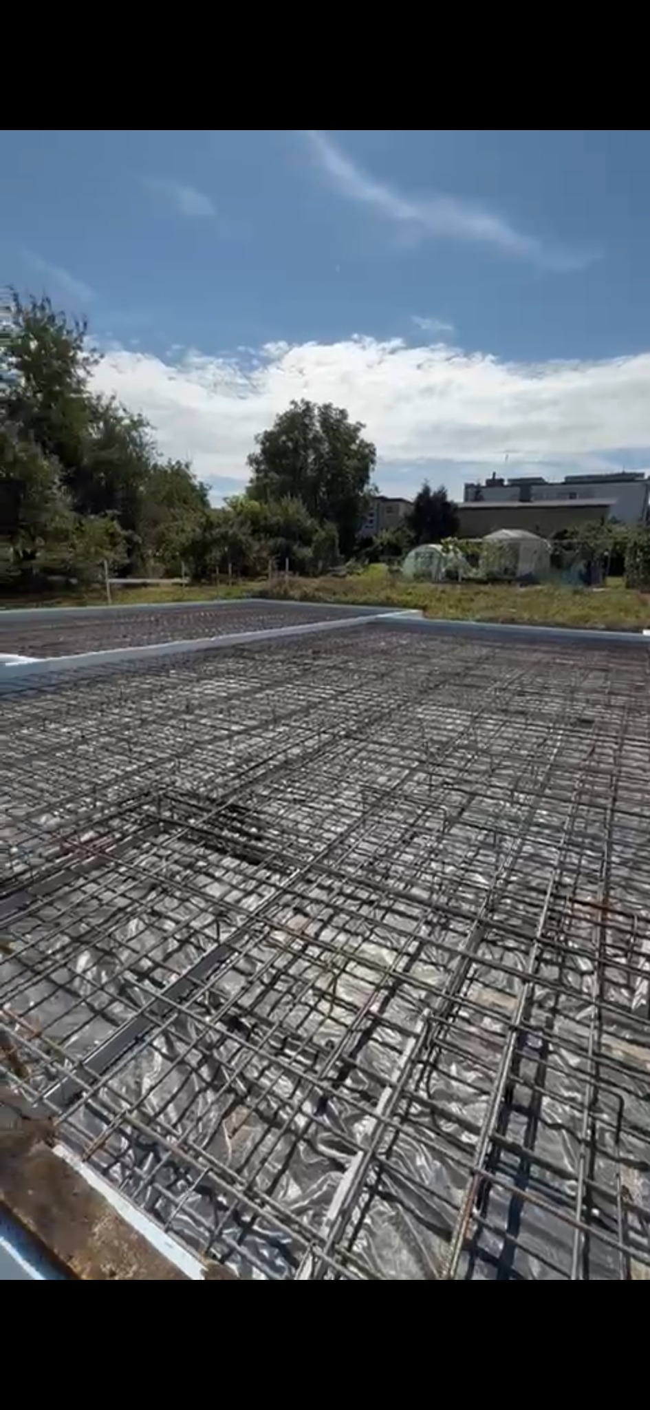 Zbrojenie płyty fundamentowej na folii izolacyjnej, widok z góry, w tle drzewa i zabudowania. Przygotowanie do wylewki betonowej, słoneczny dzień.