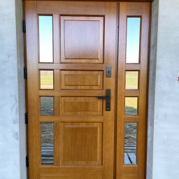 MONTERIA GRZEGORZ STAWINOGA - Drewniane drzwi zewnętrzne z naświetlem bocznym i szybami, widoczne na tle surowego muru w trakcie wykończenia, osadzone na drewnianym podeście.