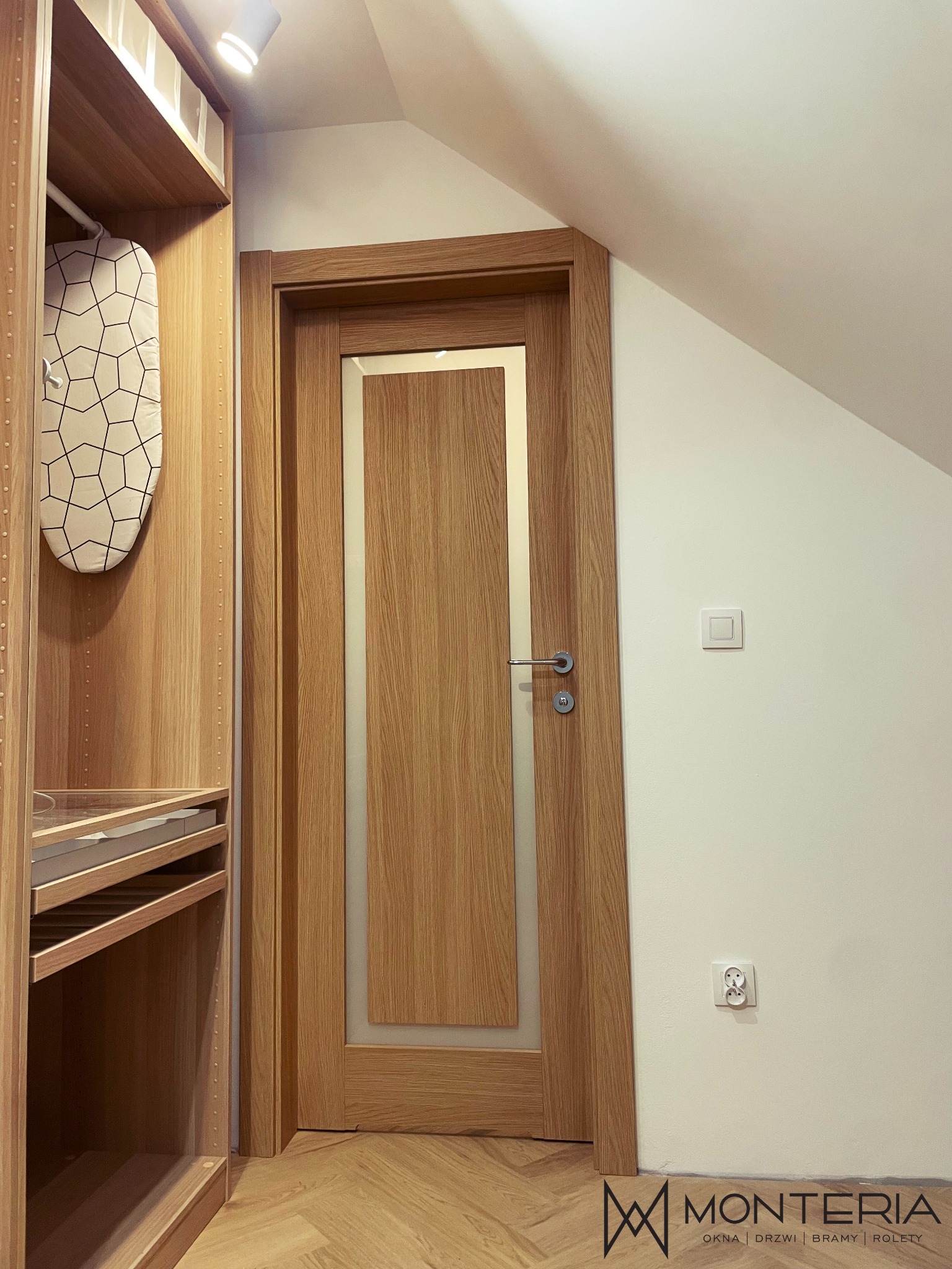 Drzwi wewnętrzne z drewnianą ramą i centralnym panelem z matowego szkła, zamontowane w pomieszczeniu ze skosami i drewnianą podłogą w jodełkę, obok wbudowana szafa z deską do prasowania.