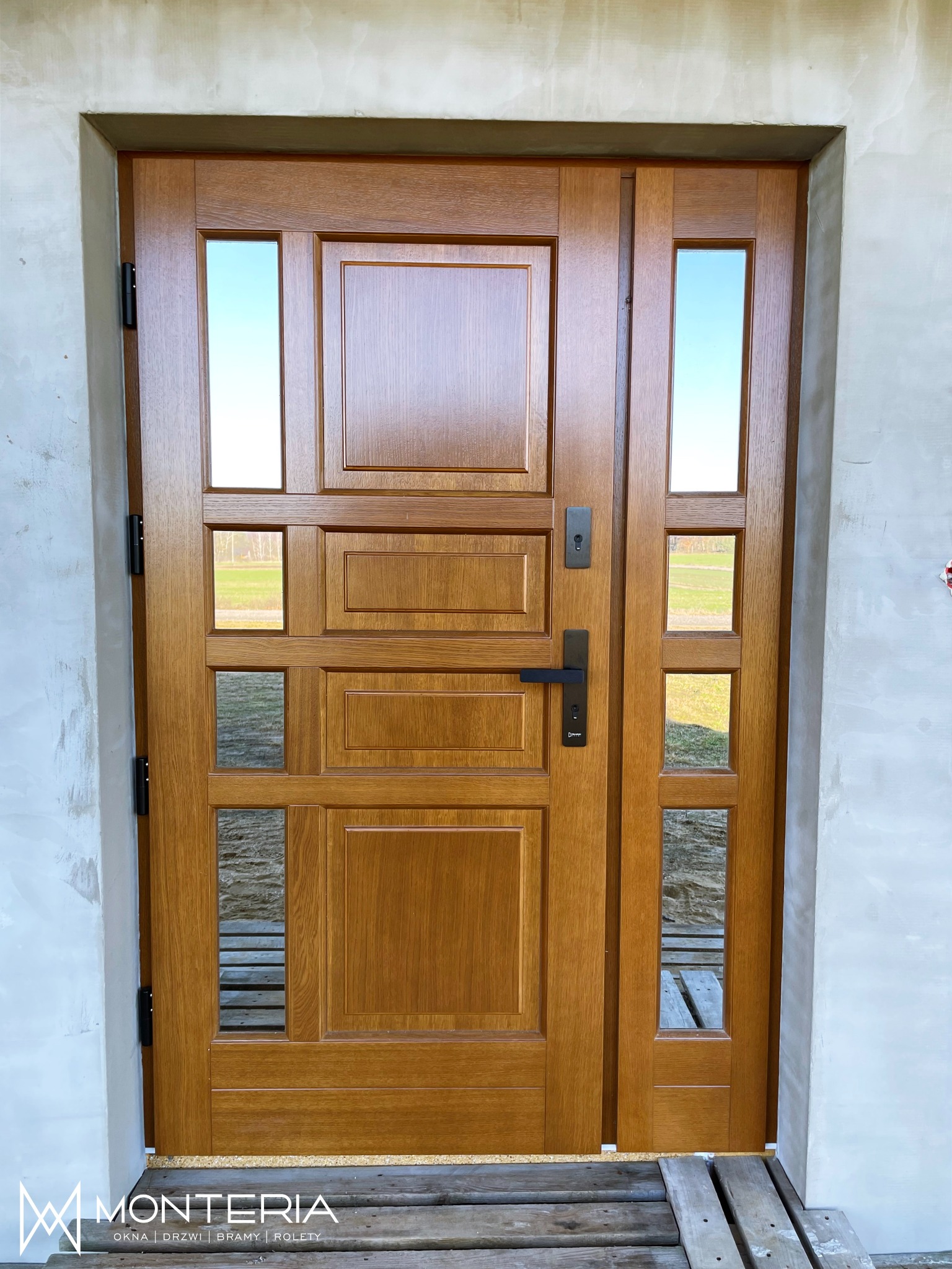 Drewniane drzwi zewnętrzne z naświetlem bocznym i szybami, widoczne na tle surowego muru w trakcie wykończenia, osadzone na drewnianym podeście.