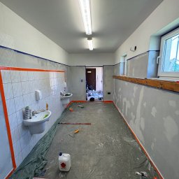 BUDEX Viacheslav Sirotin - Wnętrze w trakcie malowania: zabezpieczone folią podłogi, taśmy malarskie na ścianach, widoczne narzędzia malarskie i niedomalowane ściany z widocznymi łatami gładzi.