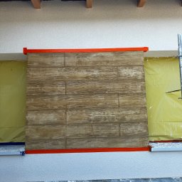 BUDEX Viacheslav Sirotin - Fragment elewacji z drewnopodobną okładziną, zabezpieczony folią i taśmą malarską, widoczny ze rusztowania. Detal wykończenia ściany zewnętrznej.