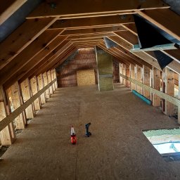 BUDEX Viacheslav Sirotin - Poddasze w trakcie adaptacji: drewniana konstrukcja dachu, płyty OSB na podłodze, narzędzia i napój. Widoczne ocieplenie i elementy instalacji. Ujęcie z dołu.