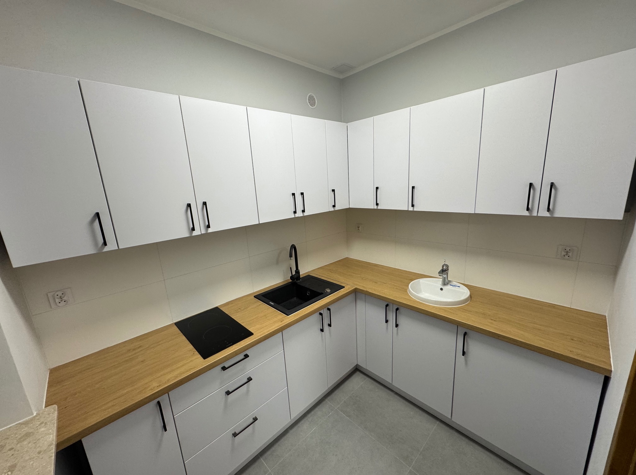 Nowoczesna, jasna kuchnia z białymi szafkami i drewnianym blatem. Czarny zlew i płyta indukcyjna. Minimalistyczne uchwyty. Świeżo po remoncie.