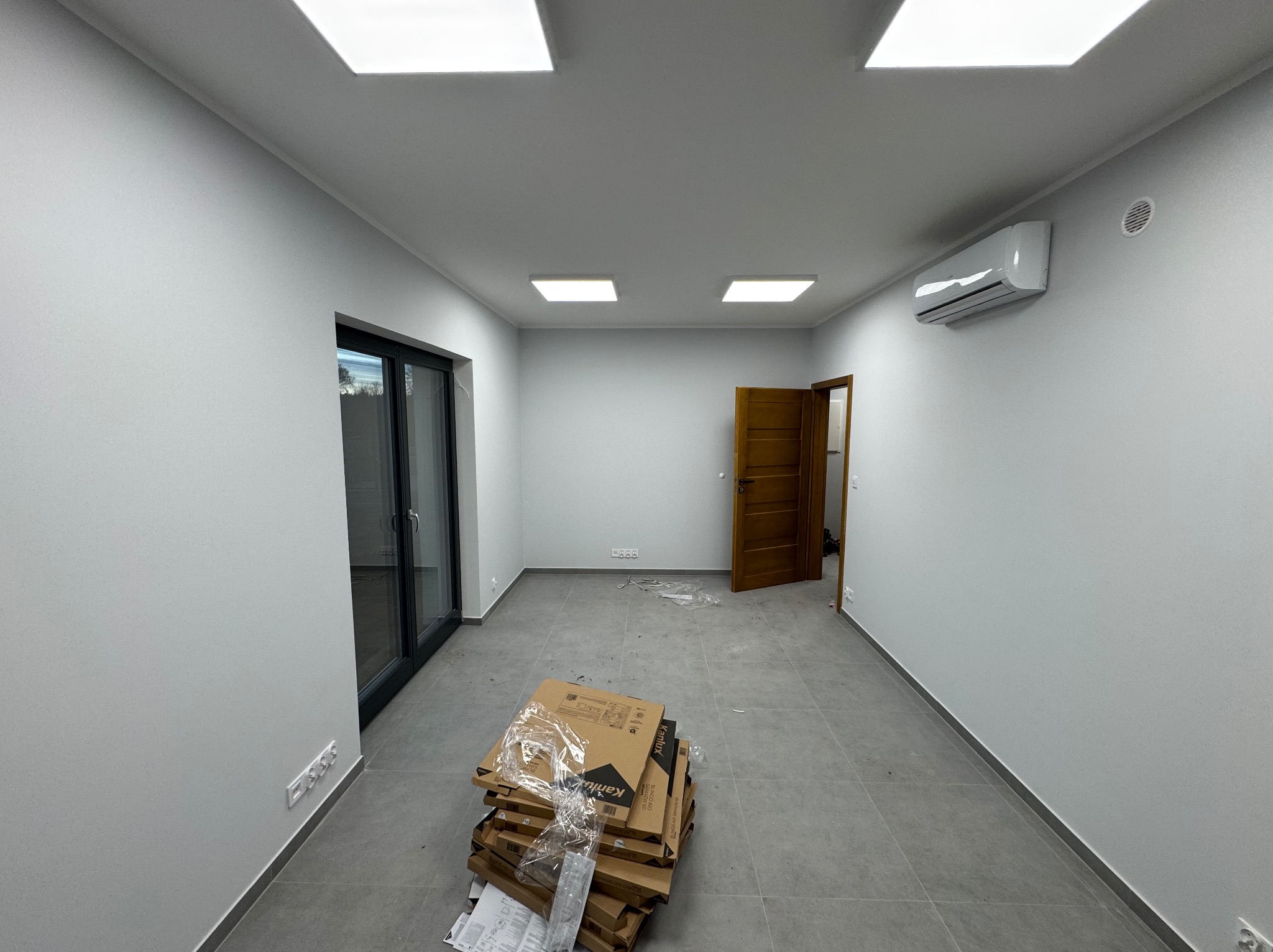Wnętrze po remoncie: świeżo pomalowane ściany na biało, szare płytki podłogowe, klimatyzacja, drzwi wewnętrzne i okno balkonowe. Na podłodze sterta kartonów.