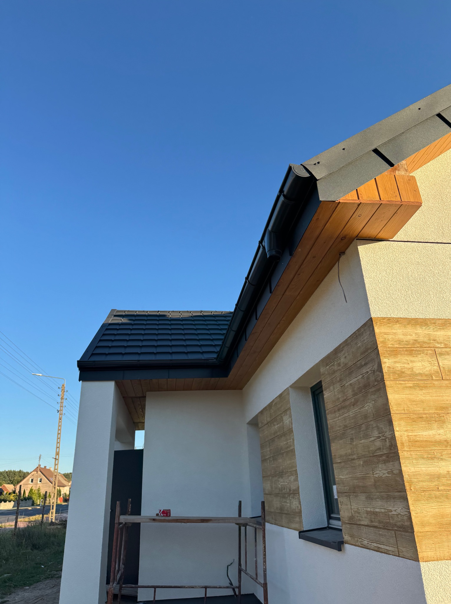 Elewacja domu z drewnianą okładziną i czarnym dachem, widziana z dołu pod kątem. Widoczne rusztowanie i fragment nieba.