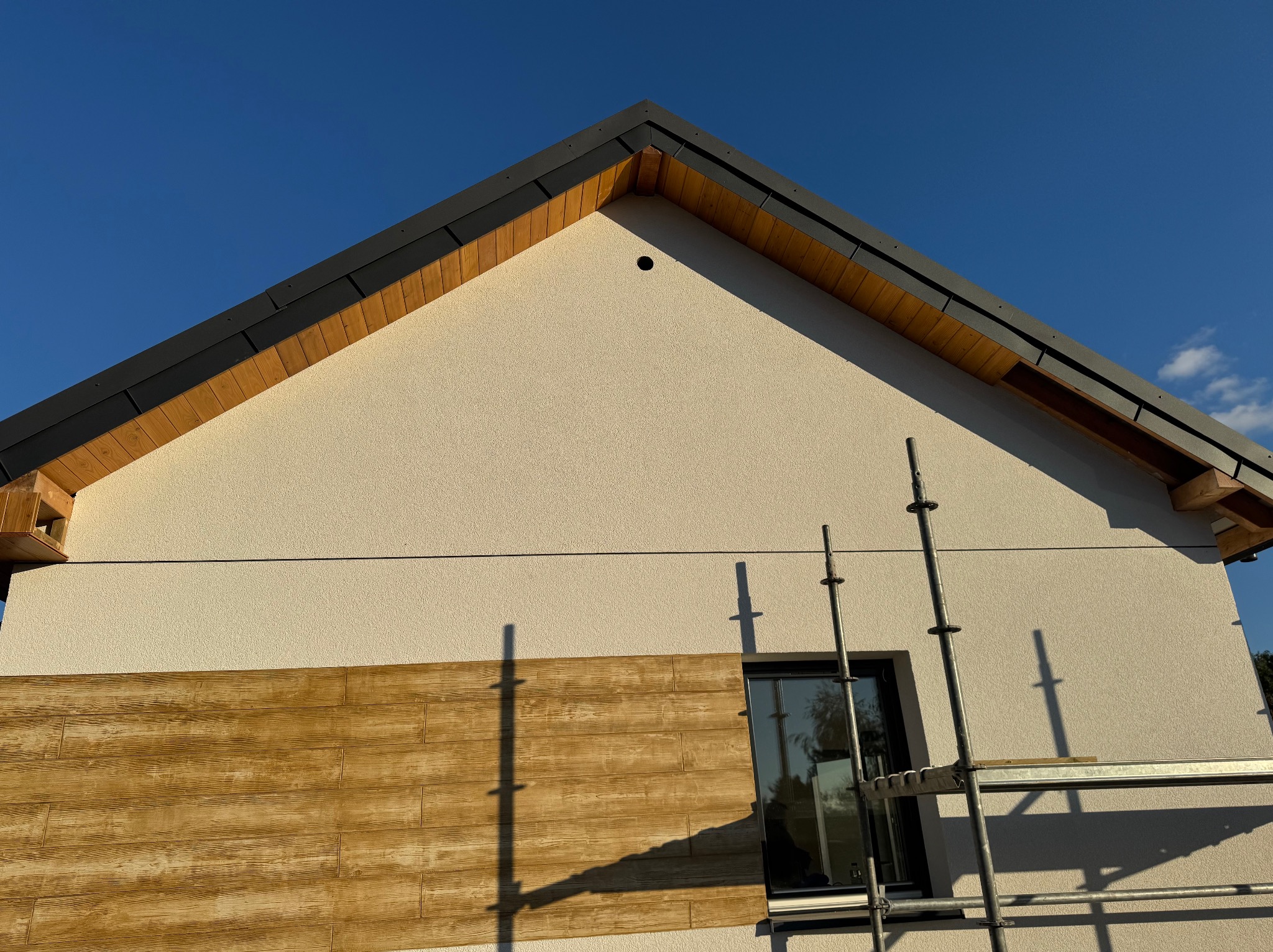 Elewacja domu w trakcie wykończenia: fragment ocieplonej ściany z dekoracyjną okładziną imitującą drewno, rusztowanie przy oknie, błękitne niebo w tle.