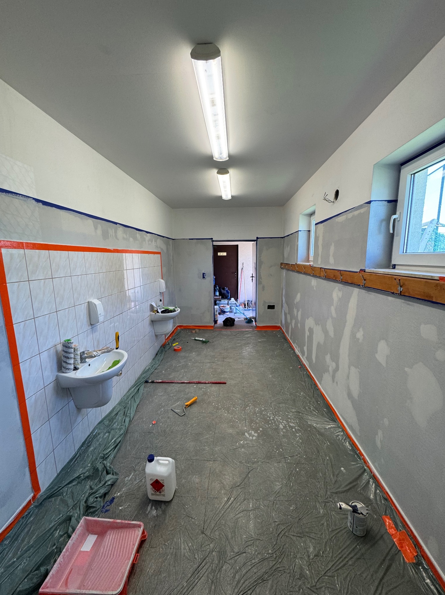 Wnętrze w trakcie malowania: zabezpieczone folią podłogi, taśmy malarskie na ścianach, widoczne narzędzia malarskie i niedomalowane ściany z widocznymi łatami gładzi.