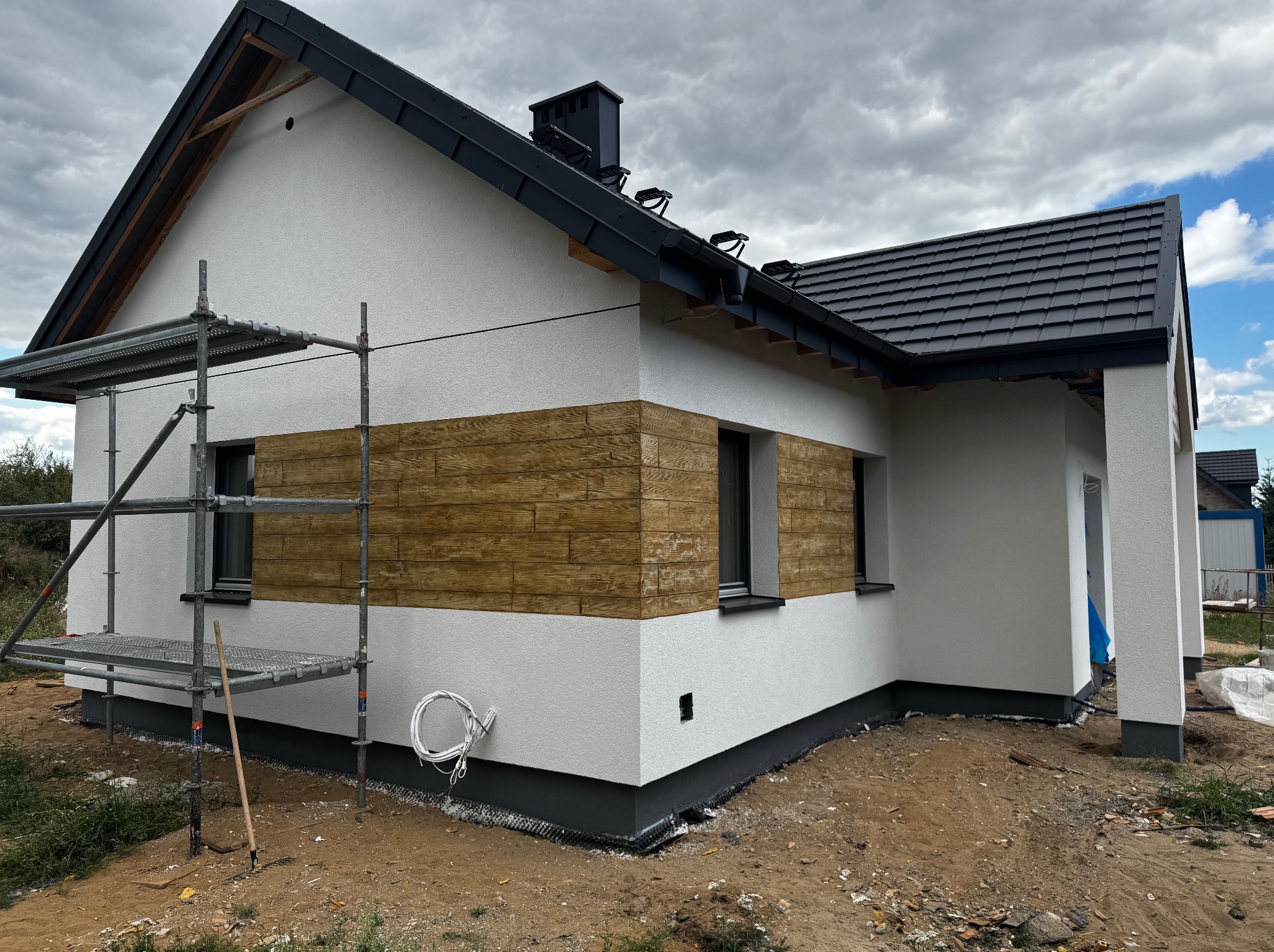 Elewacja domu jednorodzinnego z dekoracyjnym pasem imitującym drewno, widoczne rusztowanie i teren wokół budynku w trakcie prac wykończeniowych.