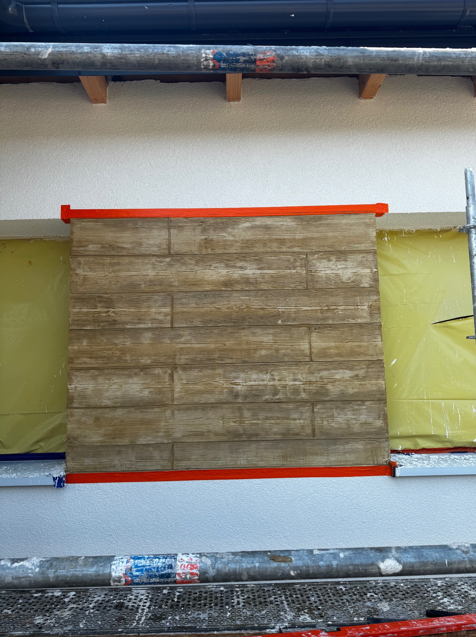 Fragment elewacji z drewnopodobną okładziną, zabezpieczony folią i taśmą malarską, widoczny ze rusztowania. Detal wykończenia ściany zewnętrznej.