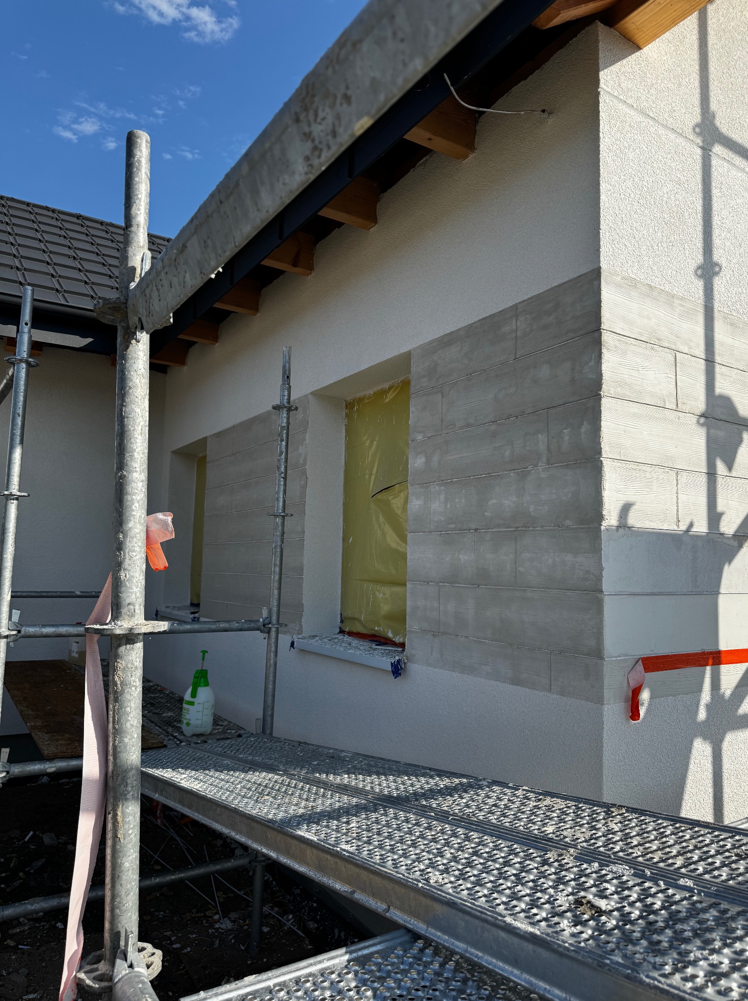 Elewacja budynku w trakcie prac wykończeniowych, widoczne rusztowanie, okna zabezpieczone folią, fragment ściany z szarą okładziną imitującą drewno.
