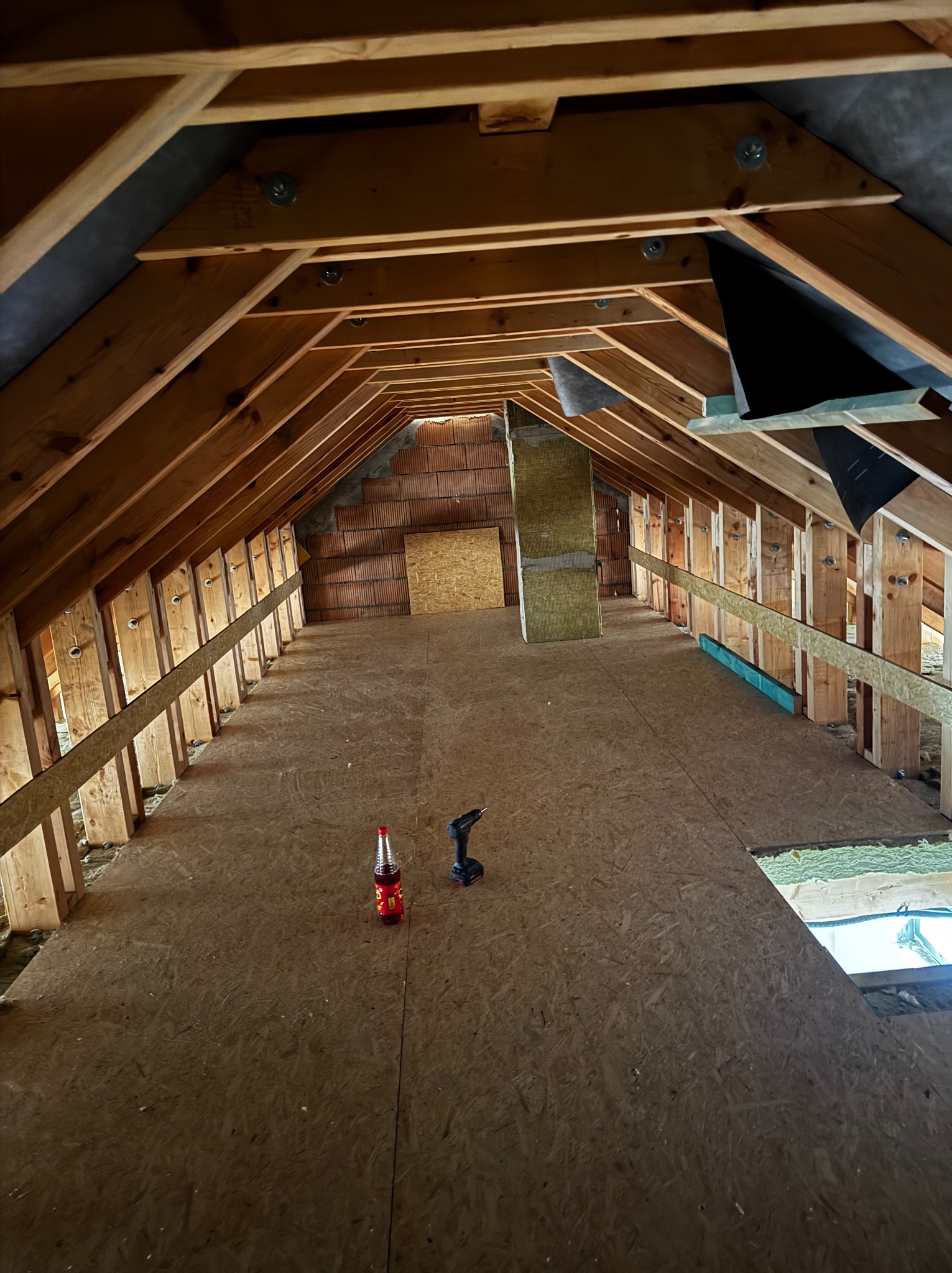 Poddasze w trakcie adaptacji: drewniana konstrukcja dachu, płyty OSB na podłodze, narzędzia i napój. Widoczne ocieplenie i elementy instalacji. Ujęcie z dołu.