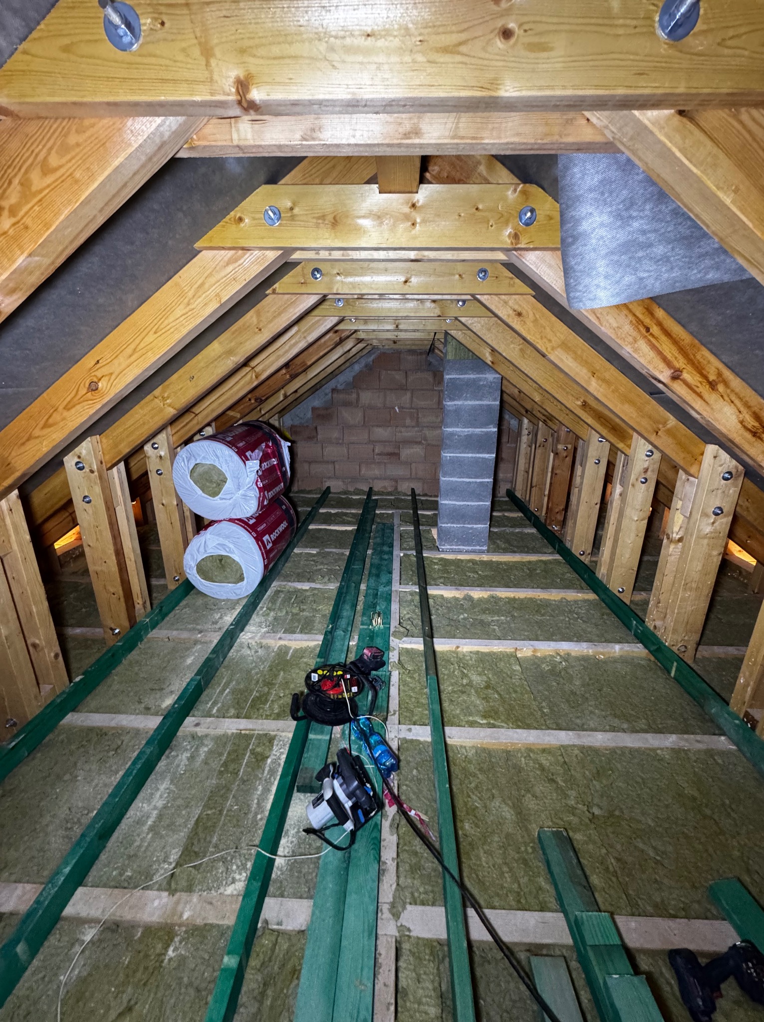 Izolacja poddasza wełną mineralną, widoczne drewniane krokwie i narzędzia. Prace remontowe w trakcie, z bliska, z perspektywy wnętrza poddasza.