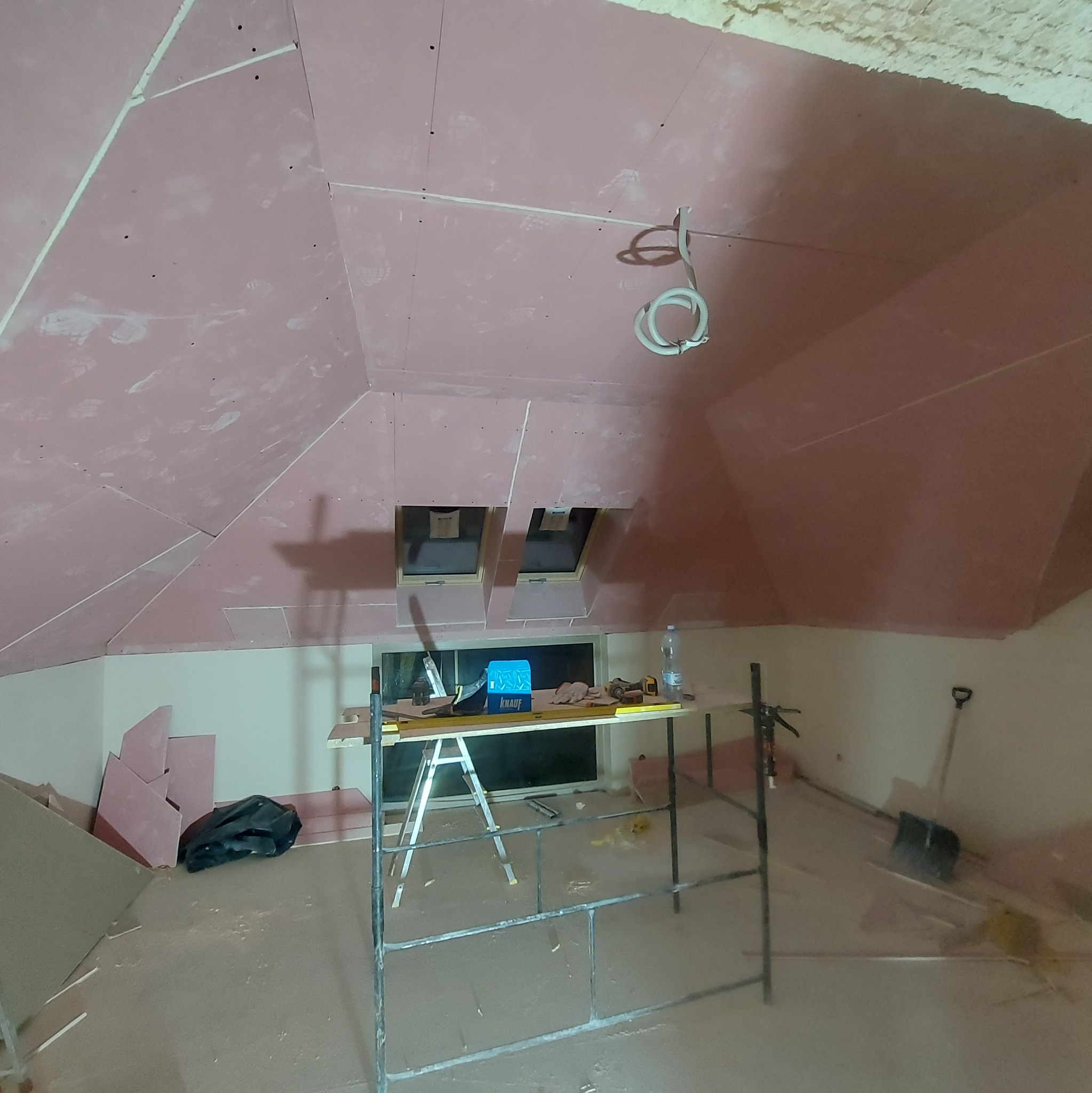 Poddasze w trakcie wykańczania płytami kartonowo-gipsowymi, widoczne okna dachowe, rusztowanie, narzędzia i materiały budowlane. Część płyt już zamontowana, inne czekają na montaż.