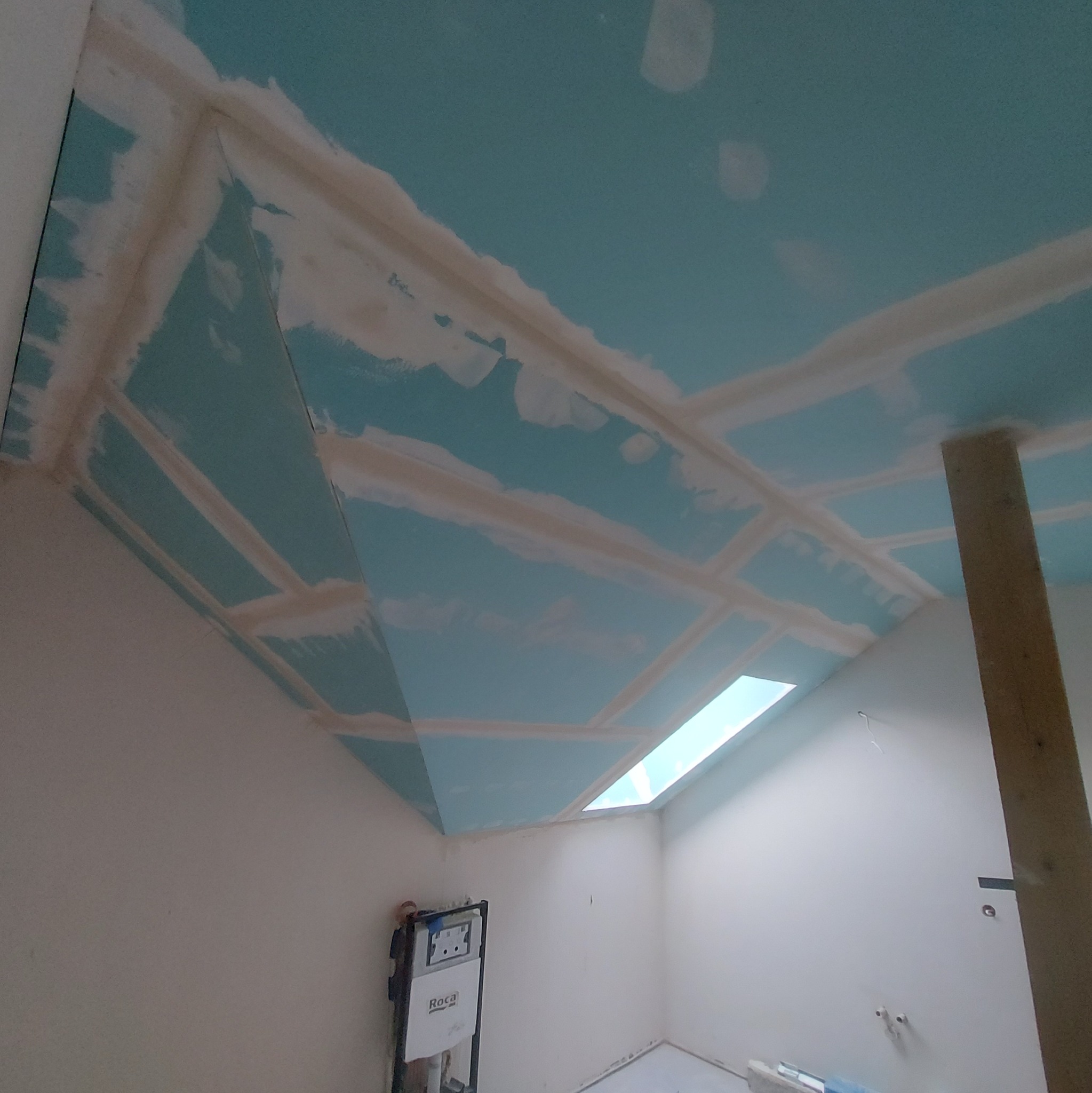 Nisko kątowe ujęcie sufitu z zamontowaną płytą kartonowo-gipsową, z widocznymi łączeniami szpachlowanymi, oświetlonego przez okno dachowe, z elementami instalacji sanitarnej na ścianie.