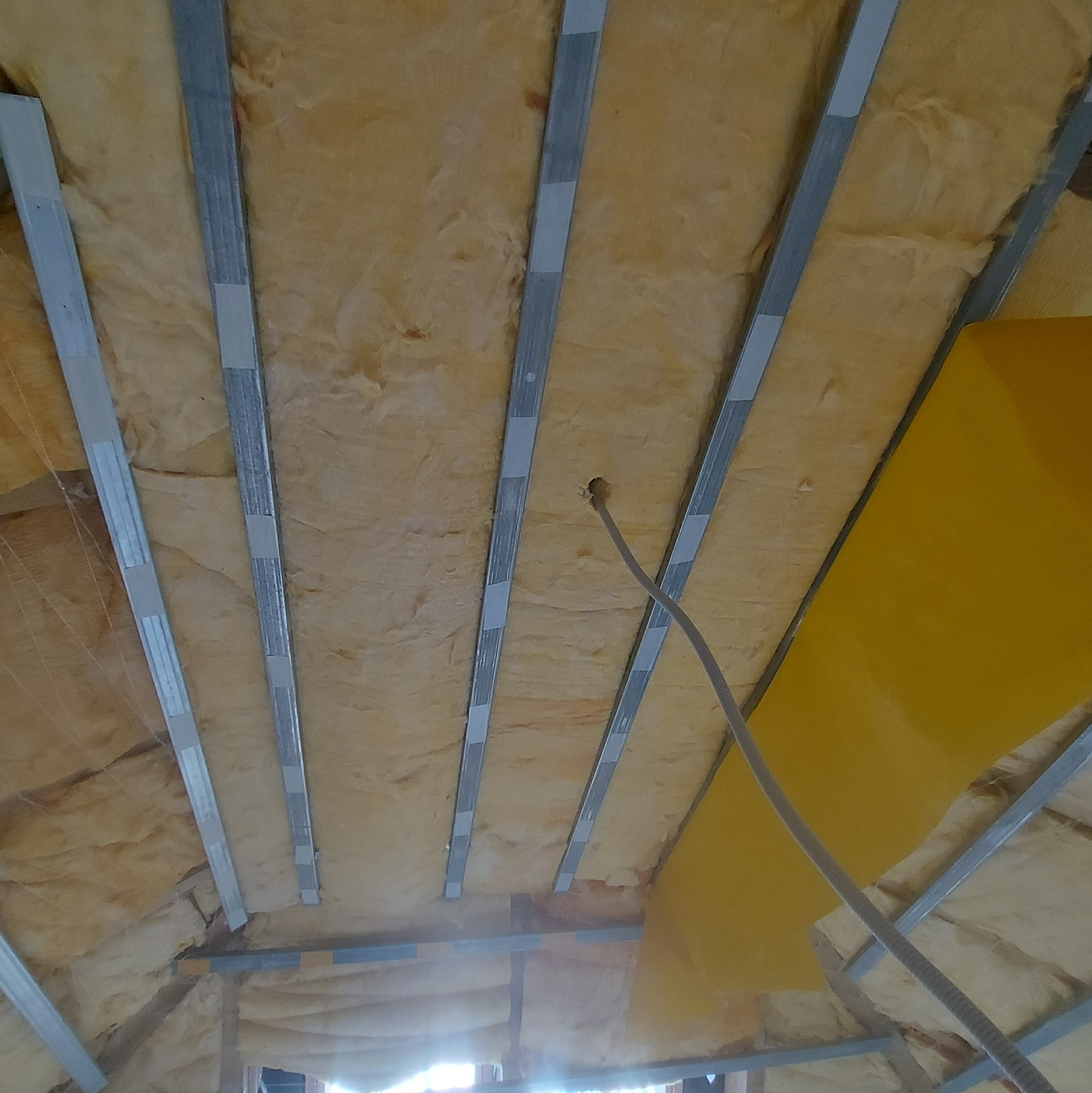 Izolacja poddasza z wełny mineralnej i metalowymi profilami, widoczny kabel elektryczny wystający z otworu w izolacji, fragment żółtej płyty.