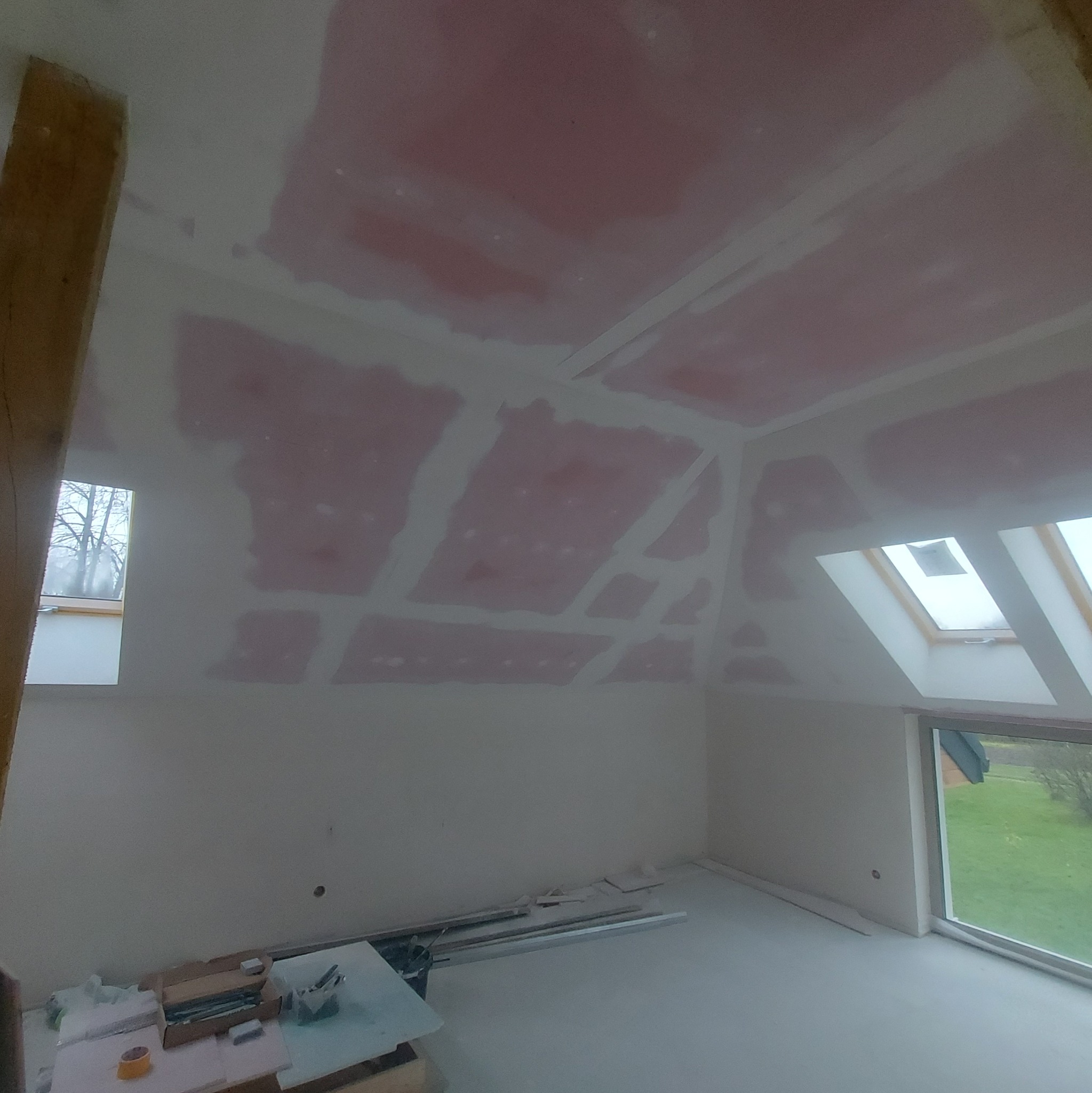 Poddasze w trakcie wykańczania, widoczne płyty karton-gips na skosach sufitu i ścian, częściowo zaszpachlowane łączenia, okno dachowe i okno w ścianie szczytowej, narzędzia na podłodze.