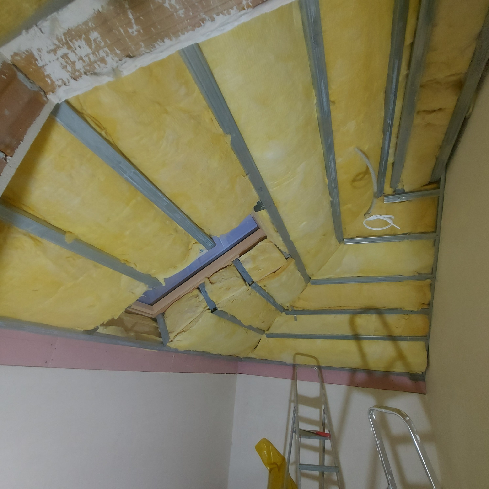 Izolacja poddasza wełną mineralną, widoczne profile metalowe, fragment okna dachowego i drabina oparta o ścianę.