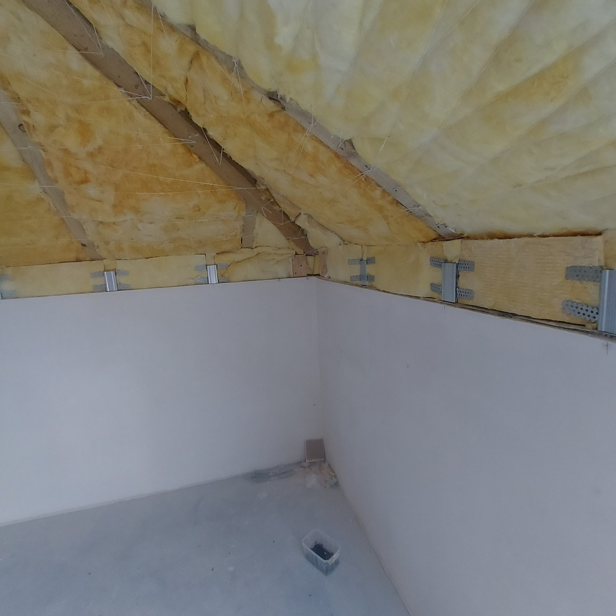 Izolacja poddasza wełną mineralną, widoczne metalowe mocowania i fragment ściany z płyt kartonowo-gipsowych, u dołu pojemnik z wkrętami.