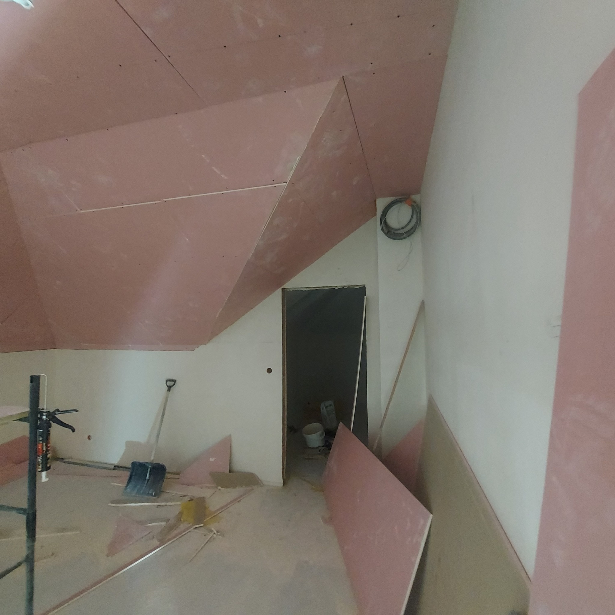 Poddasze w trakcie wykańczania płytami kartonowo-gipsowymi, widoczne narzędzia, resztki materiałów i otwór drzwiowy prowadzący do kolejnego pomieszczenia.