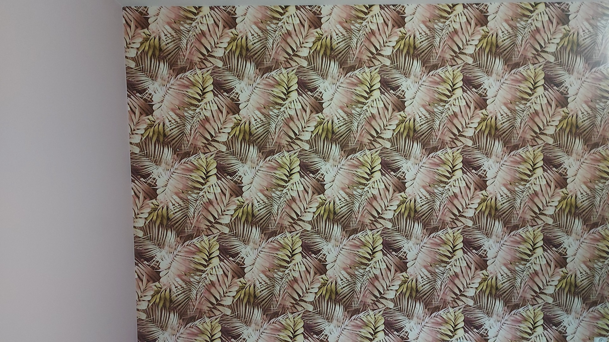 Ściana pokryta tapetą w tropikalny wzór z motywem liści palmowych w odcieniach brązu, beżu i różu, widoczna z bliska, z fragmentem gładkiej, jasnej ściany po lewej stronie.