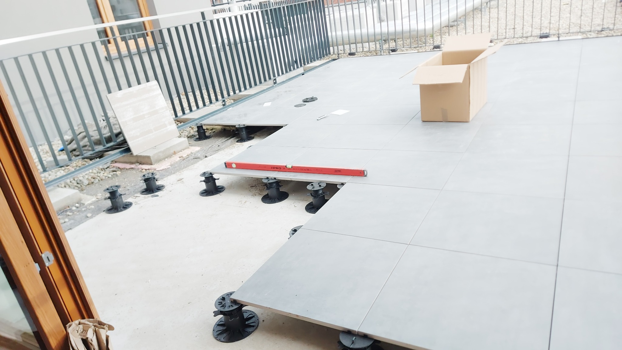 Montaż szarych płyt tarasowych na regulowanych wspornikach, poziomica na płytach, otwarte kartonowe pudło, balustrada w tle, etap prac.