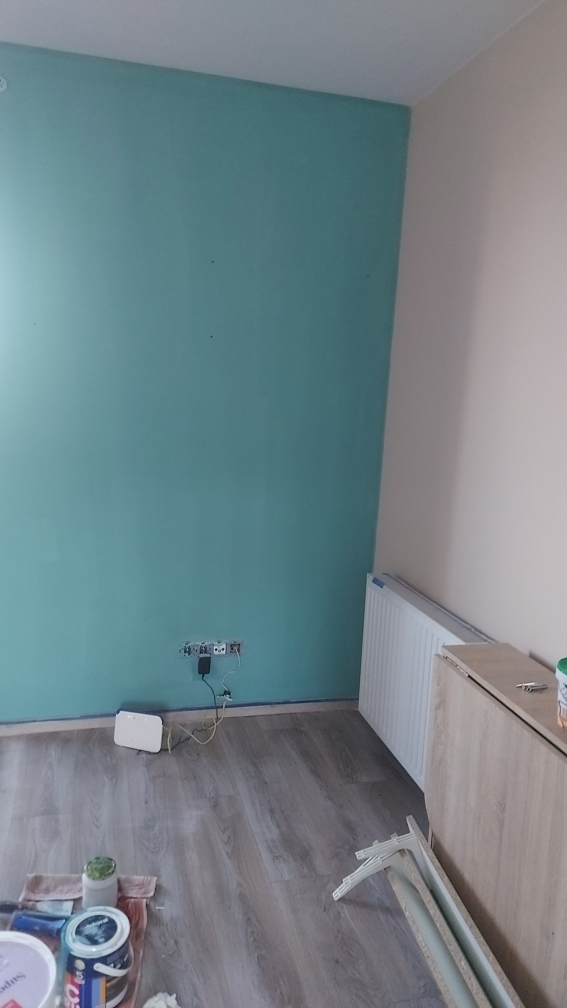 Świeżo pomalowana ściana w odcieniu turkusu z widocznymi puszkami farb i akcesoriami malarskimi na podłodze z paneli, gniazdka elektryczne bez osłon.