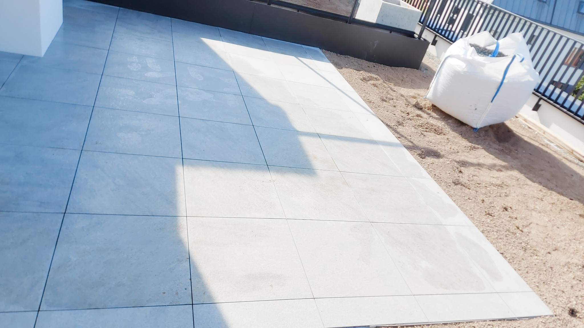 Ułożona jasnoszara podłoga z płytek na tarasie, obok duży worek z piaskiem i widok na balustradę.