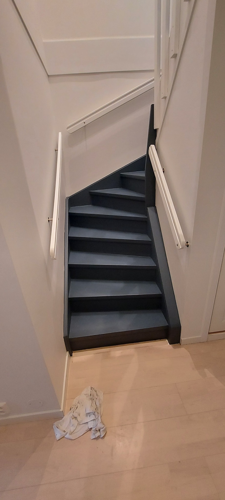 Pomalowane na ciemno schody z białymi poręczami w jasnym wnętrzu, na podłodze leży brudna szmatka.