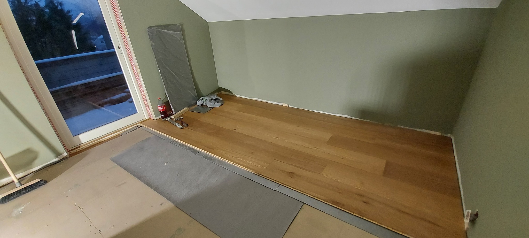Fragment pokoju z oknem balkonowym w trakcie układania drewnianej podłogi, widoczne narzędzia, butelka napoju i folia ochronna przy ścianie pomalowanej na oliwkowy kolor.
