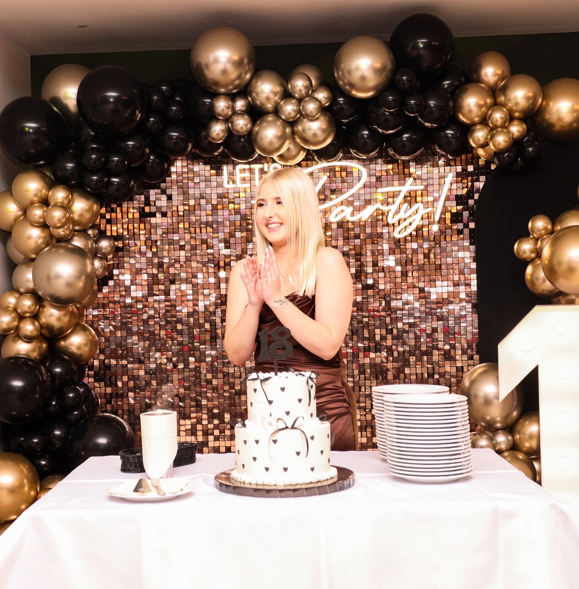 Uśmiechnięta blondynka klaszcze przed tortem urodzinowym z dekoracją '18' na tle błyszczącej ścianki i balonów w kolorach złota i czerni. Dekoracja urodzinowa.