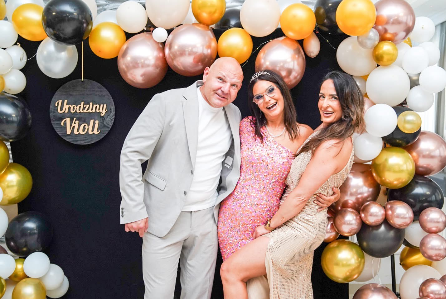 Uśmiechnięte osoby na tle dekoracji balonowych z napisem 'Urodziny Violi'. Kobiety w cekinowych sukienkach, mężczyzna w garniturze. Radosna atmosfera.