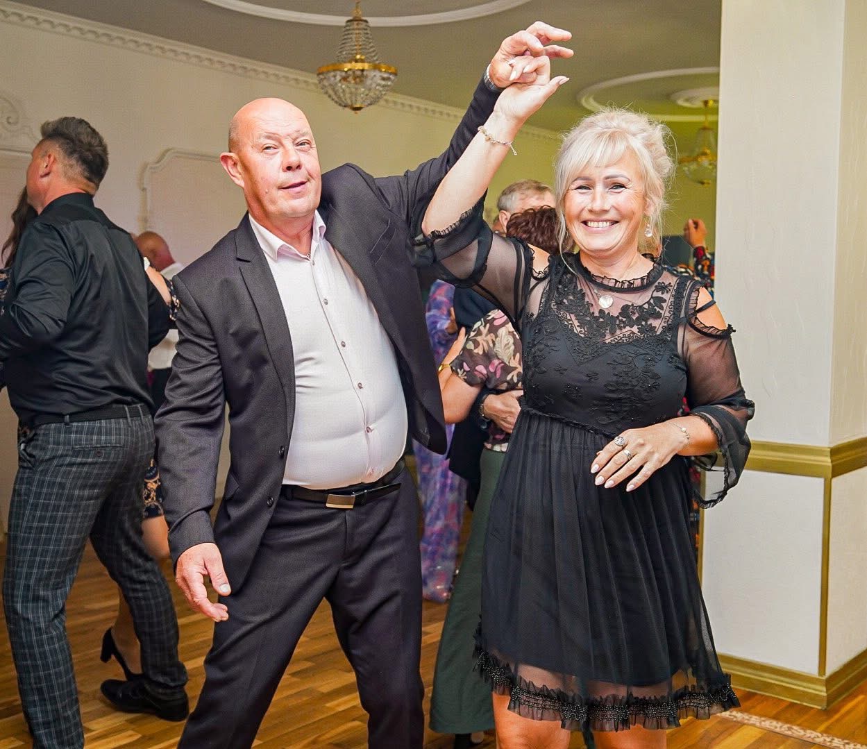 Para w tańcu na parkiecie podczas uroczystości. Kobieta w czarnej sukience, mężczyzna w garniturze. W tle widoczni inni goście i elementy wystroju sali.