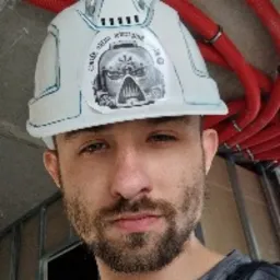 Hydraulik w kasku z naklejką na tle czerwonych rur instalacyjnych. Widoczne elementy konstrukcji ściany. Ujęcie z bliska, skupione na twarzy fachowca.