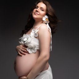 Pulina Semeniuk Photography - Uśmiechnięta, ciemnowłosa kobieta w ciąży, ubrana w białą sukienkę z kwiatowymi aplikacjami, pozuje na ciemnoszarym tle, z rękami ułożonymi na brzuchu i kwiatem we włosach.