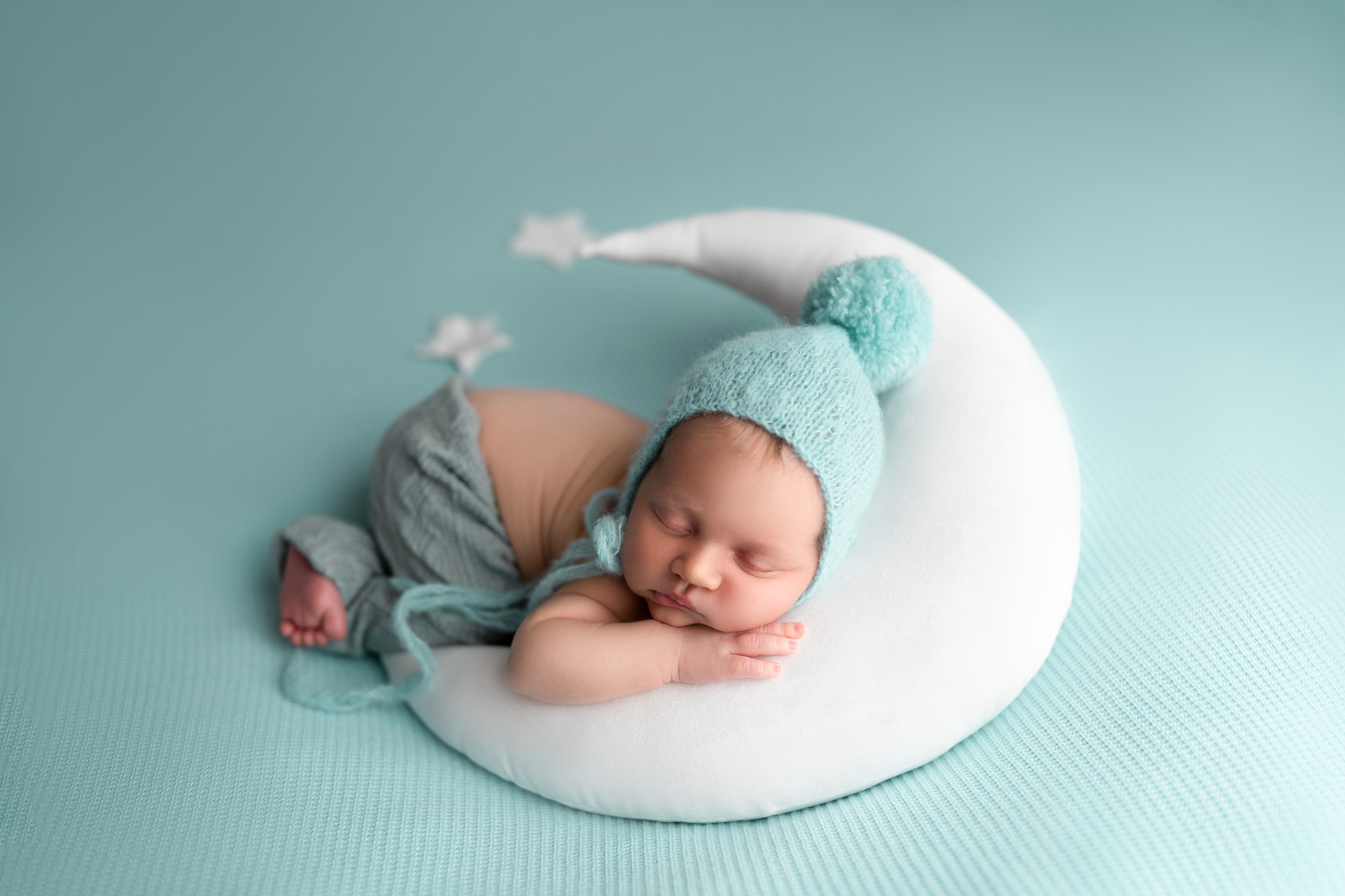 Śpiący noworodek w turkusowym czapeczce z pomponem, ułożony na białej poduszce w kształcie księżyca z gwiazdkami na turkusowym tle.