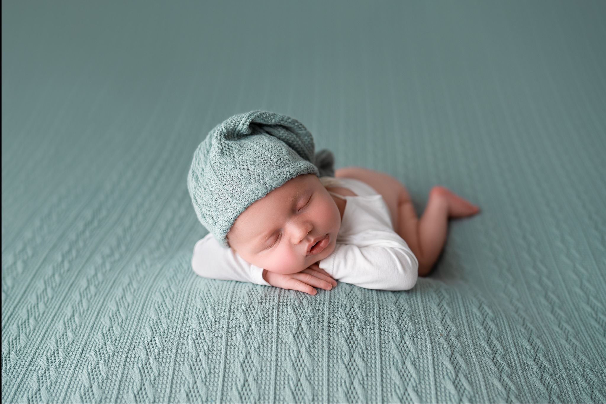 Śpiący noworodek w błękitnej czapce, leżący na brzuchu na teksturowanym, błękitnym kocyku.
